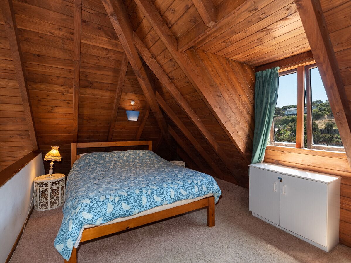 Un dormitorio con techo de madera, una cama con colcha azul estampada, una lámpara de noche, un armario blanco y una ventana con vistas a la vegetación.