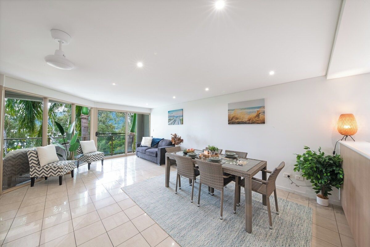 A spacious living area with a dining table, sofas, and a large window offering a view of greenery.