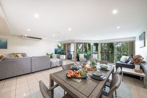 A modern living room with a dining area, grey sofas, a large window, and a flat-screen TV. - Phil's Place on Airlie (Airlie Beach)