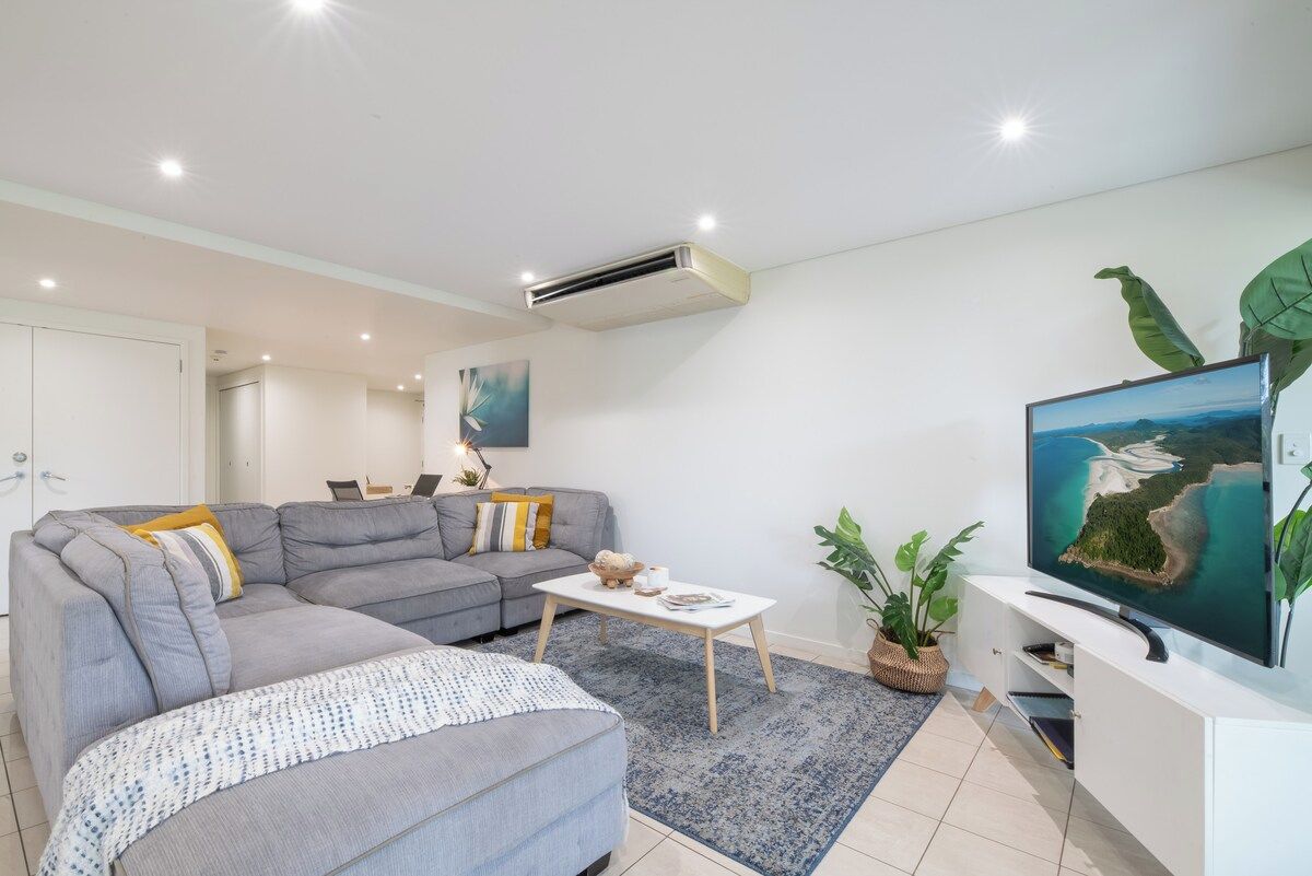 Een moderne woonkamer met een grijze hoekbank, een salontafel, een flatscreen televisie en een groot raam.