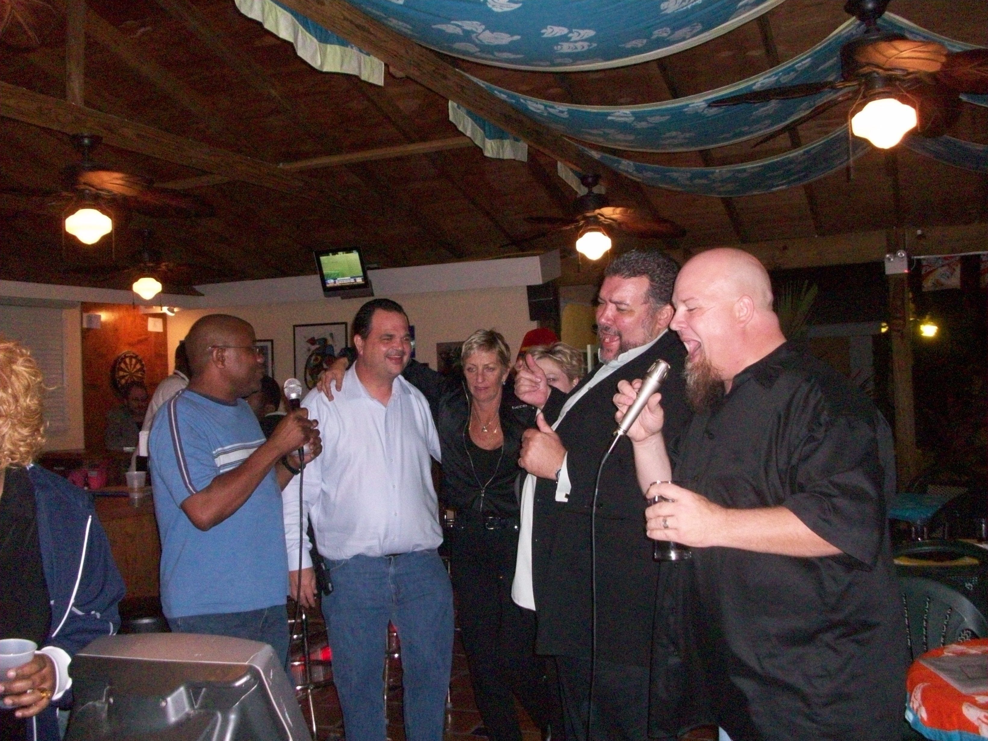 A group of people in a dimly lit room with wooden beams on the ceiling and a television mounted on the wall.