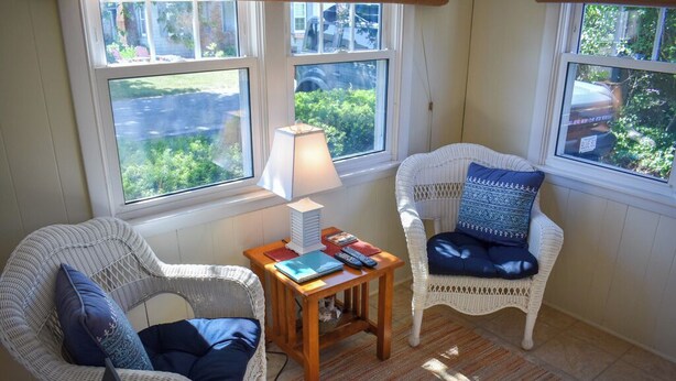 Een gezellige kamer met rieten stoelen, een klein houten tafeltje met boeken en een lamp, en grote ramen met uitzicht op groen buiten.
