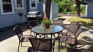 Een terras met een ronde glazen tafel, rieten stoelen, een barbecue en een potplant.