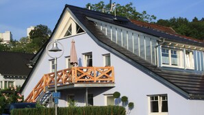 Una casa con techo inclinado, un balcón con barandillas de madera y una campana en un mástil.