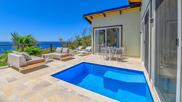 A modern outdoor pool area with a view of the sea, furnished with lounge chairs and a dining table. - Villa Topaz Guest Villa Above West Bay With 180 Degree Views @ Villa Topaz (West Bay)
