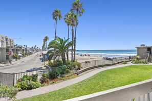A coastal street with a view of the beach, palm trees, and a clear blue sky.