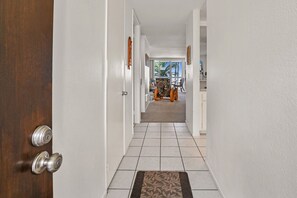 A hallway with a wooden door, white walls, and a tiled floor leading to a room with a chair and a table. - Charming Upper Beachfront Cottage-Breathtaking Ocean Views-D8 North Coast Village (Oceanside)