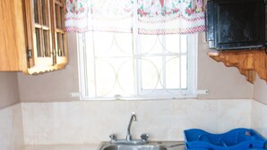 A kitchen with wooden cabinets, a stainless steel sink, and a window with a decorative curtain. - Belle Air Paradise JM (Runaway Bay)
