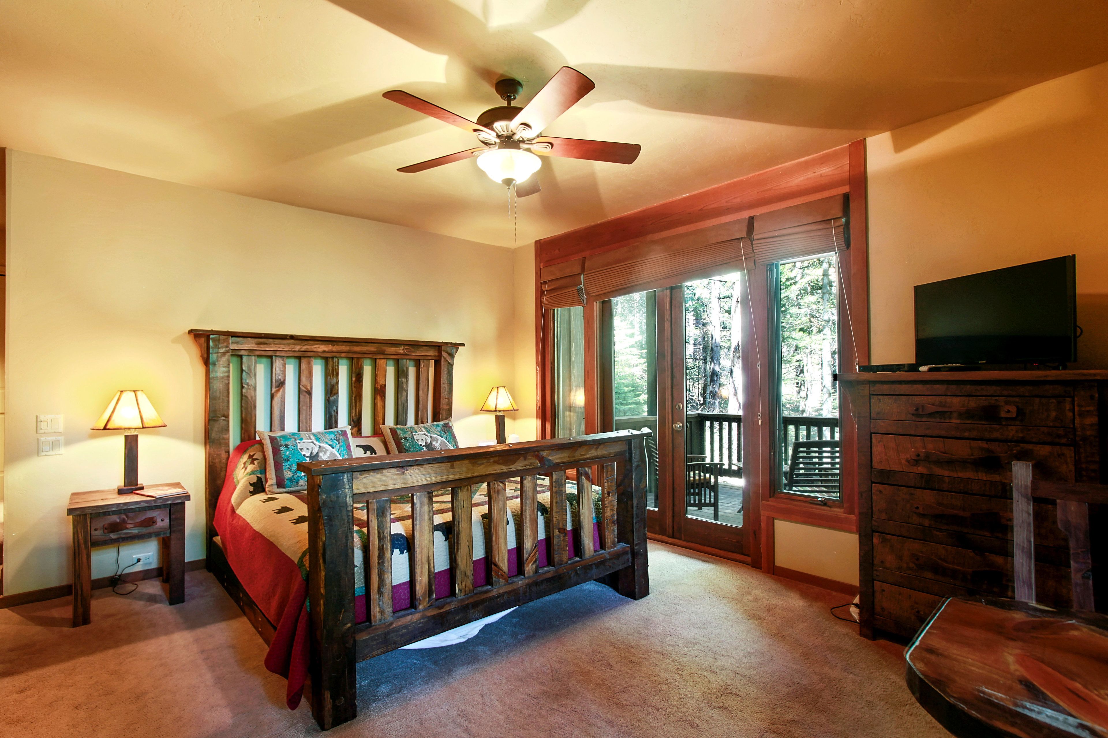 A bedroom with a wooden bed, two bedside lamps, a ceiling fan, a television, and a large window with a view of trees.