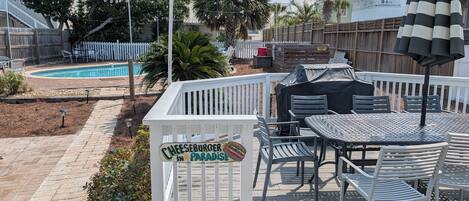 Una terraza con un cartel que dice 'CHEESEBURGER IN PARADISE', muebles de exterior y una piscina al fondo.