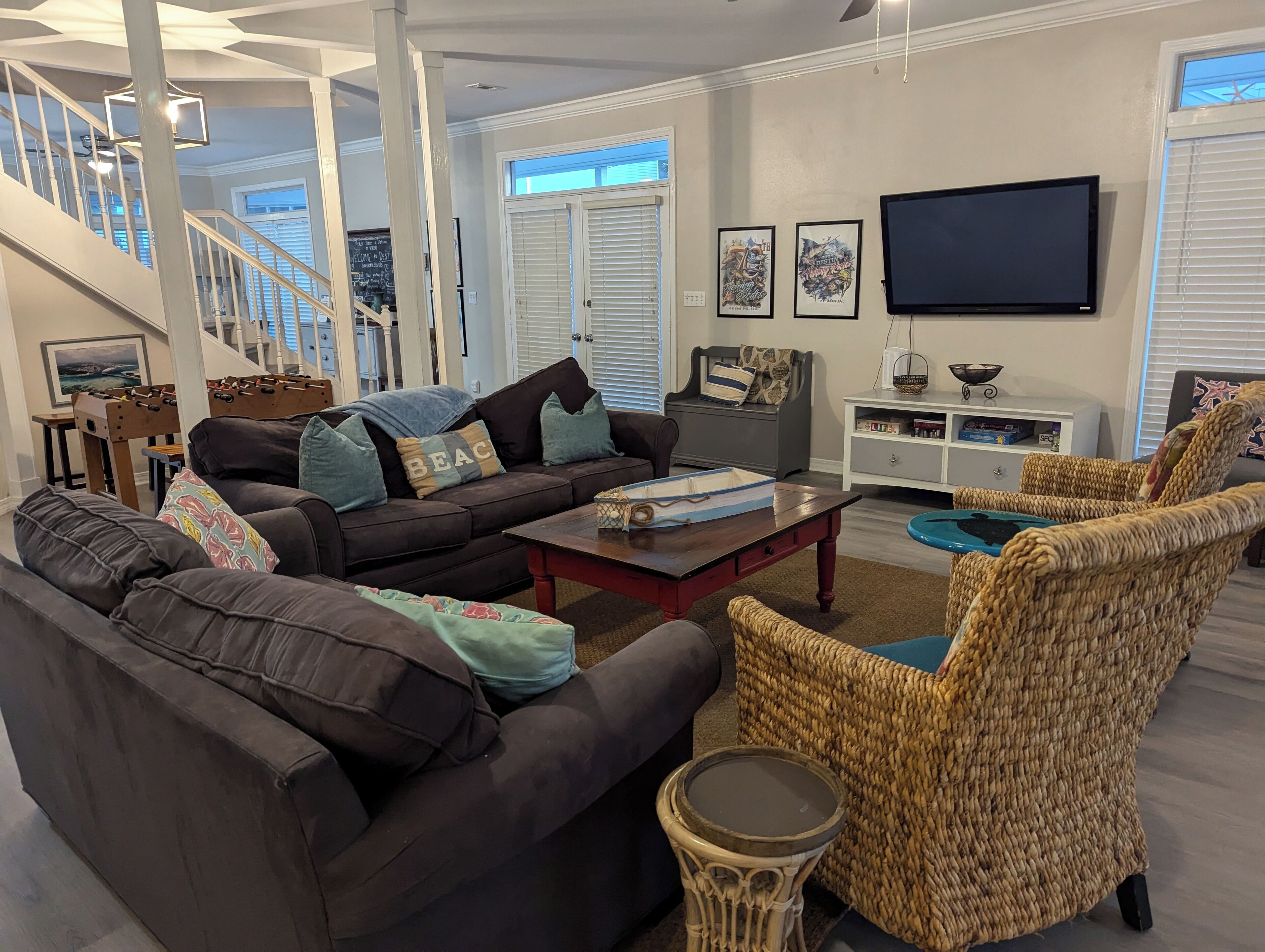 A spacious living room with a dark brown sofa, a wicker chair, and a coffee table. A staircase with a railing is visible in the background.