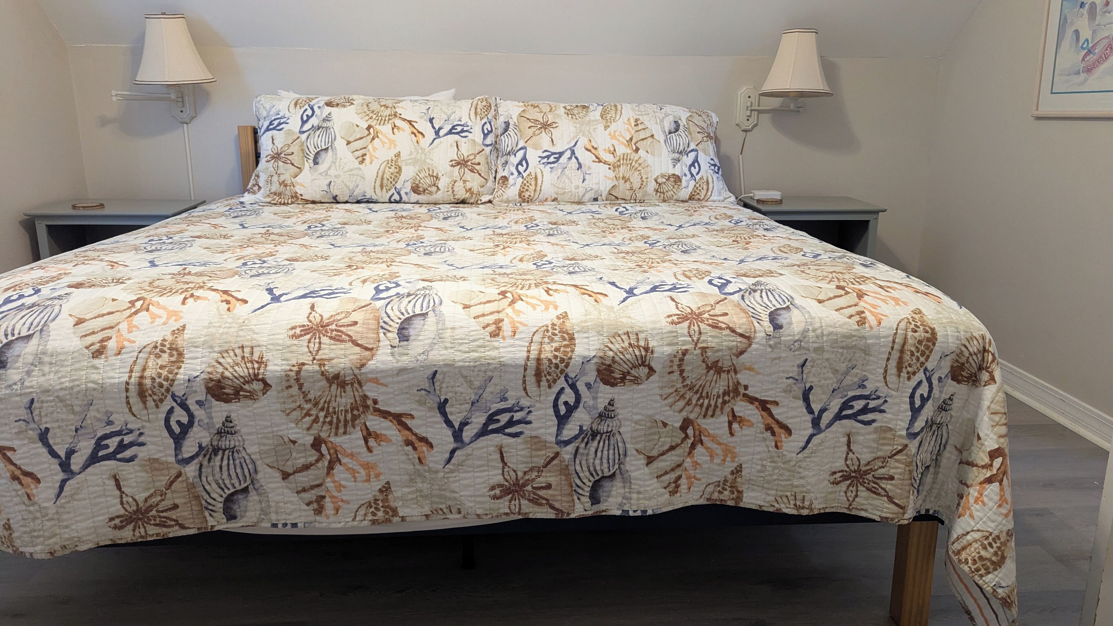 Un lit recouvert d’une couvre-lit à motifs floraux et de coquillages, agrémenté d’un coussin assorti, et flanqué de deux tables de chevet sur lesquelles sont posées des lampes.