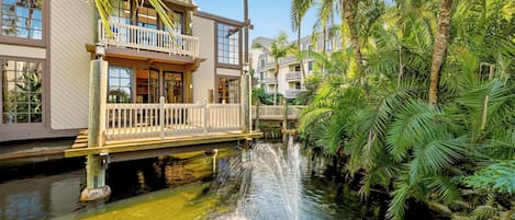 A waterfront hotel with a balcony, a water feature, and lush greenery.
