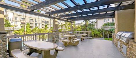 An outdoor patio with stone tables and benches, a grill, and a view of residential buildings.