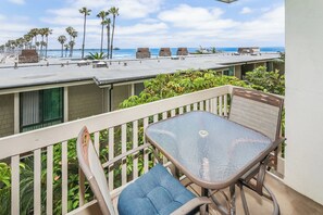 A balcony with a table and chairs, overlooking a beach with palm trees and a pier.