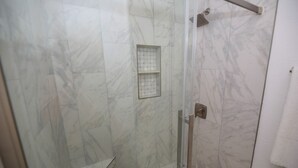 Ducha con cerramiento de vidrio, pared de mármol blanco, una ventana pequeña y un perchero para toallas.