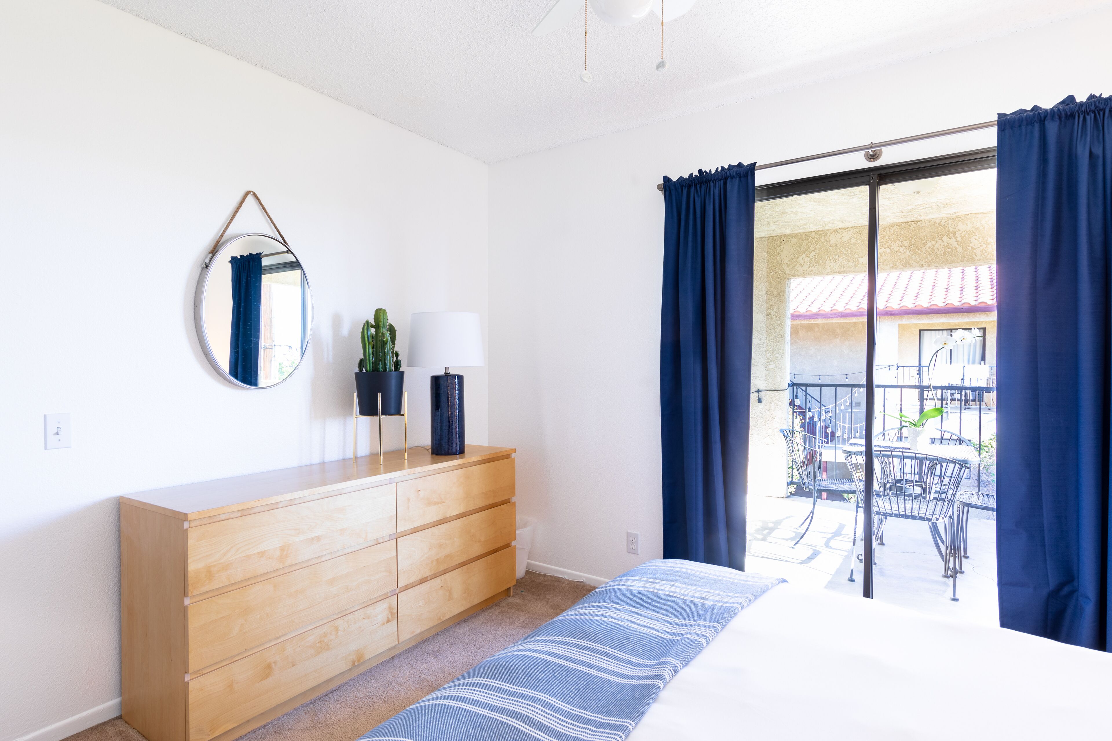 A bedroom with a wooden dresser, a bed with a striped bedspread, a round mirror, and a window with blue curtains.