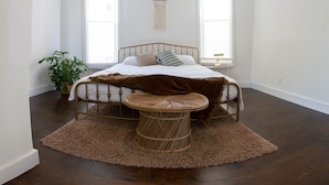 Une chambre à coucher avec un lit, une petite table d’appoint ronde en osier, une plante en pot et deux fenêtres.