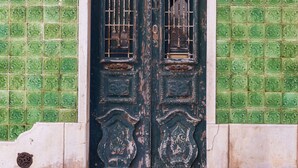 Une porte à deux battants ornée de sculptures complexes et surmontée d’une grille métallique, le tout intégré à un bâtiment dont la façade est recouverte de carreaux verts à motifs.
