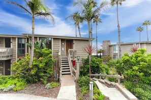 A modern building with a covered walkway leading to a door, surrounded by lush greenery and palm trees.