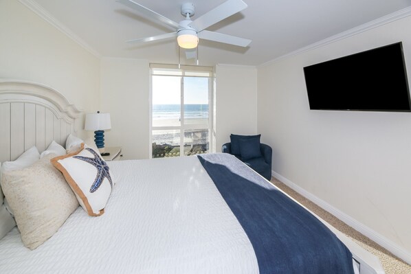 A hotel room with a bed, a TV, a ceiling fan, a window with a view of the ocean, and a chair.