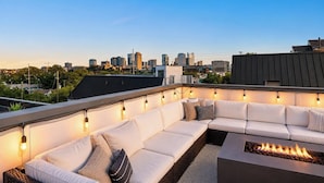 A rooftop patio with a white sofa, fire pit, and string lights. - Rooftop Style · Fire Pit · Wet Bar · Skyline Views (Nashville)