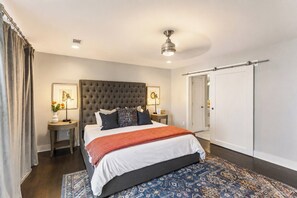 Une chambre à coucher avec une tête de lit capitonnée, un grand lit recouvert d’une couverture rouge, deux tables de chevet avec des lampes, un tapis à motifs et une porte coulissante.
