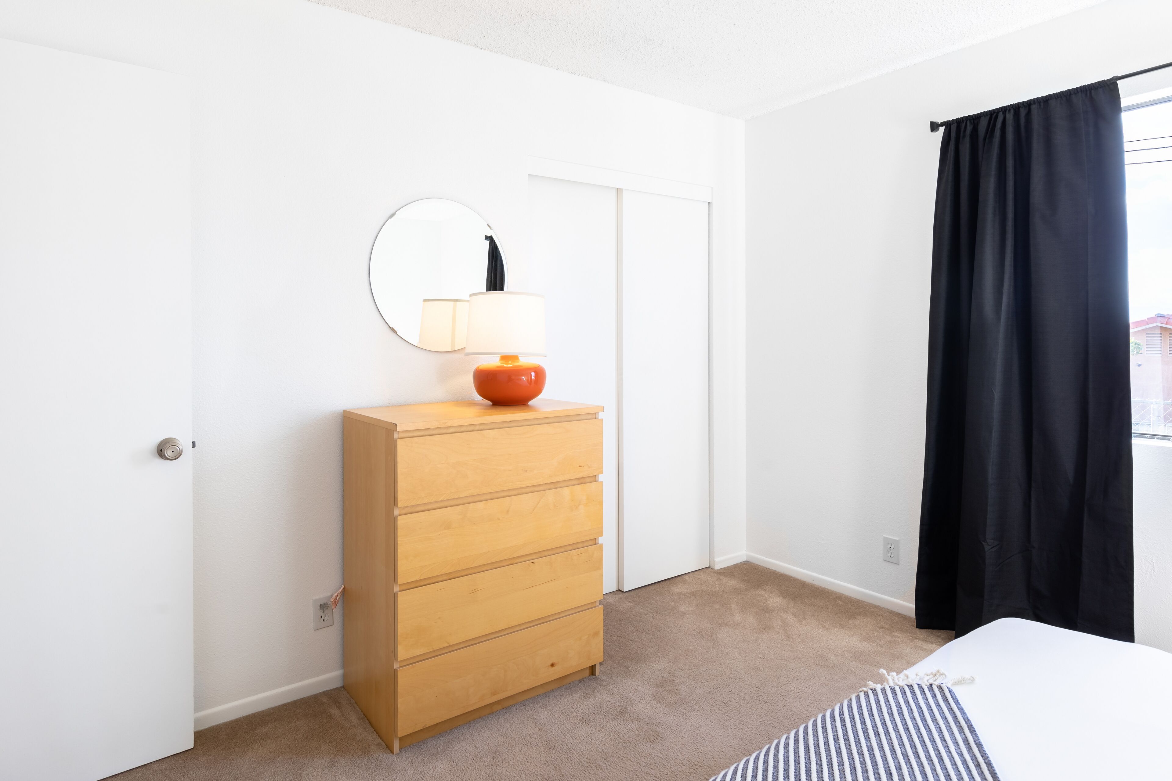 Een slaapkamer met een houten dressoir, een ronde spiegel, een bed met een gestreept dekbed en een raam met zwarte gordijnen.