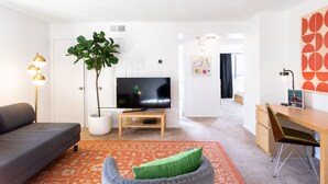 En moderne stue med en grå sofa, en grøn polstret stol, et fladskærms-tv på et træstel, en stor potteplante og et skrivebord med tilhørende stol.