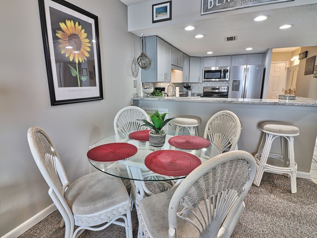 A dining area with a glass table surrounded by wicker chairs, a framed sunflower artwork, and a kitchen in the background.