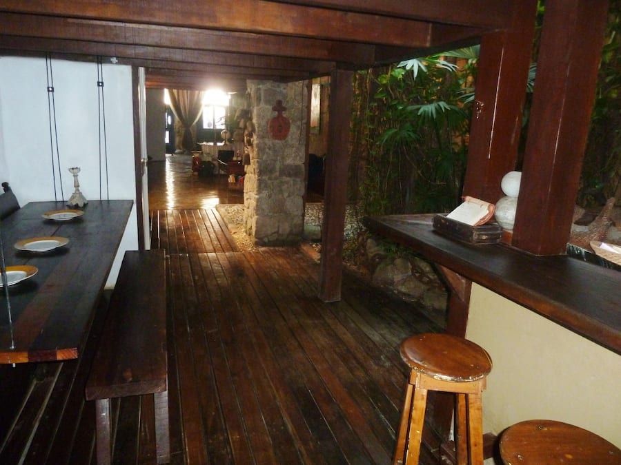 Una zona con barra de madera, taburetes, pared de piedra con vegetación y un largo banco de madera.