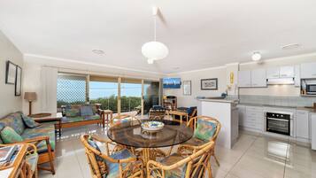 A modern living room with a glass dining table surrounded by wicker chairs, a sofa with cushions, and a kitchen area with white cabinets and appliances.