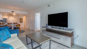 A modern living room with a flat-screen TV, a sofa, a coffee table, and a dining area in the background.