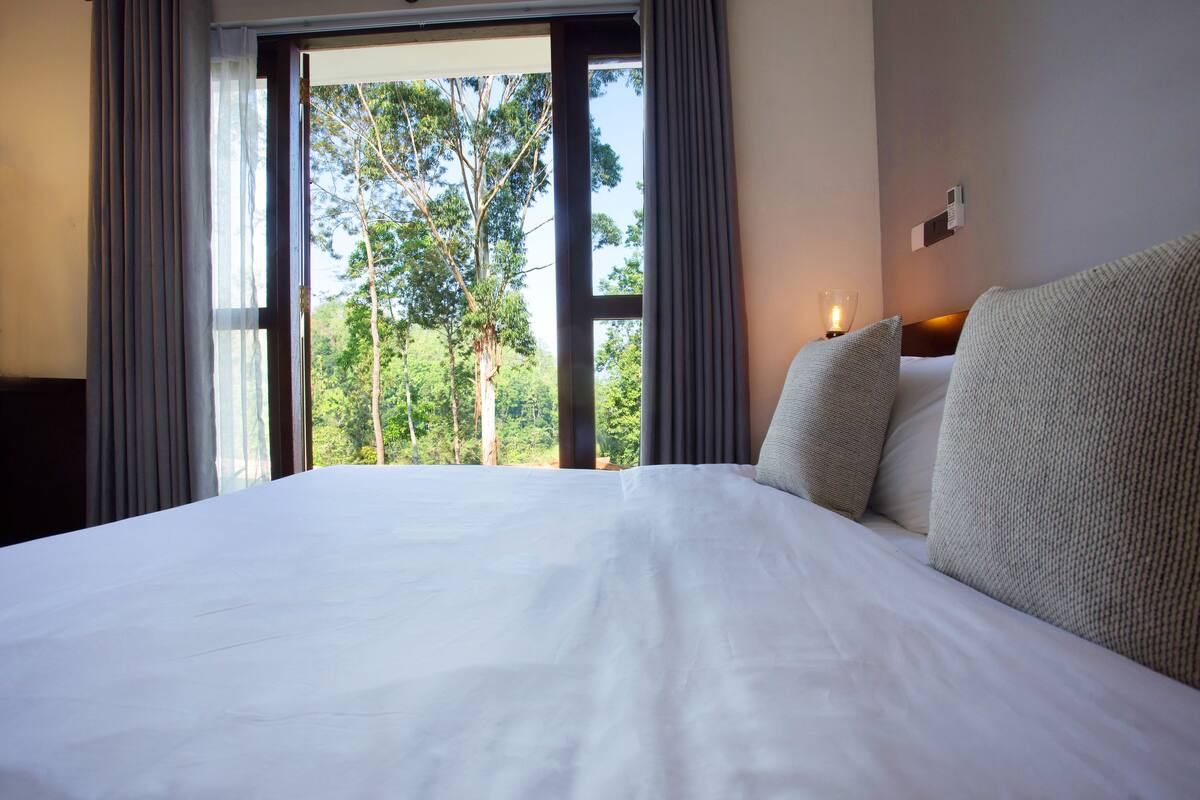 A large bed with white linens, a view of trees through a window, and a wall-mounted thermostat.