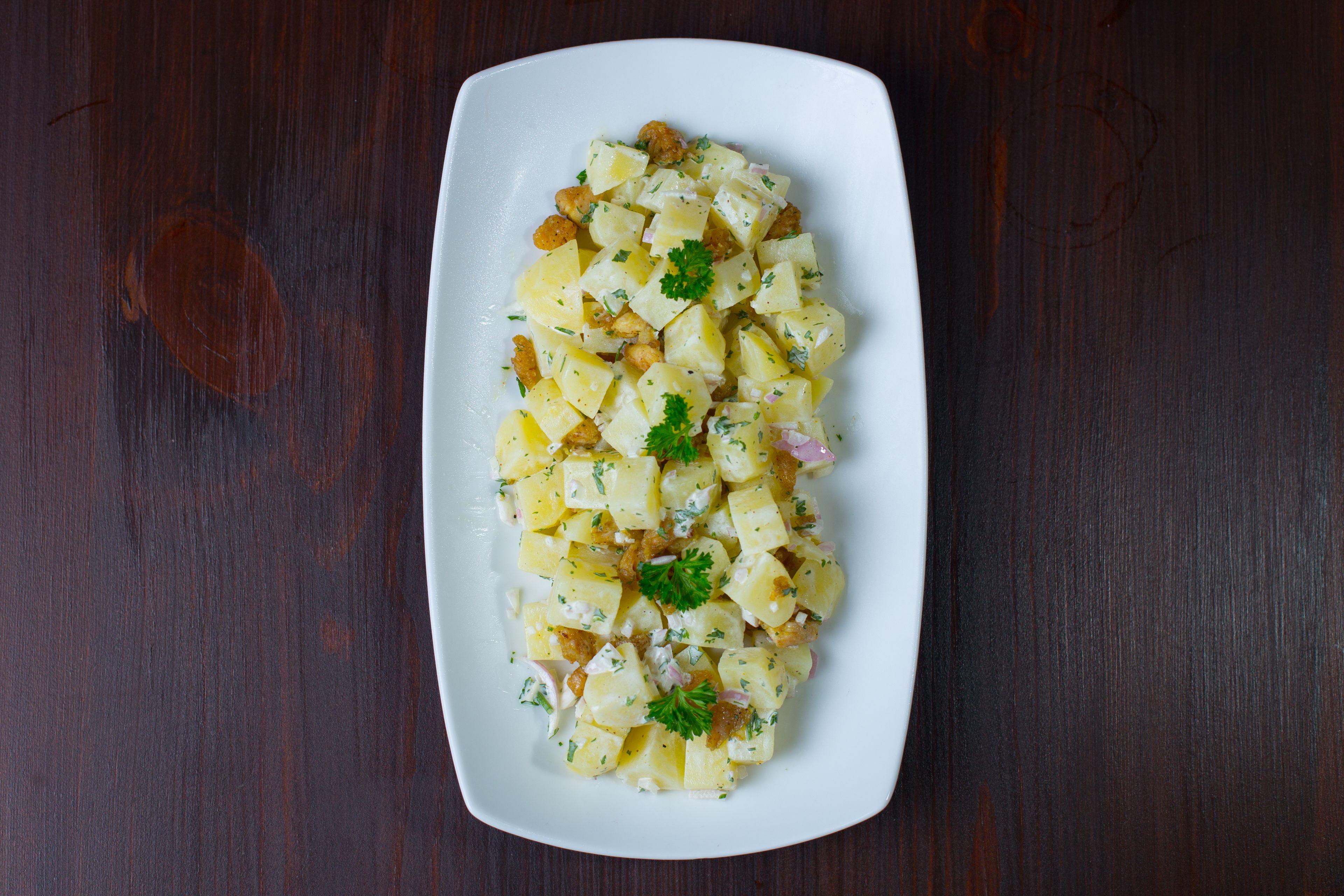 A white rectangular plate with a potato salad on a dark wooden table.