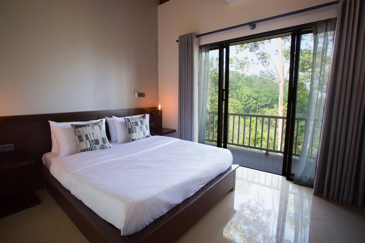 A hotel room with a large bed, a nightstand, a lamp, and a balcony with a view of trees.
