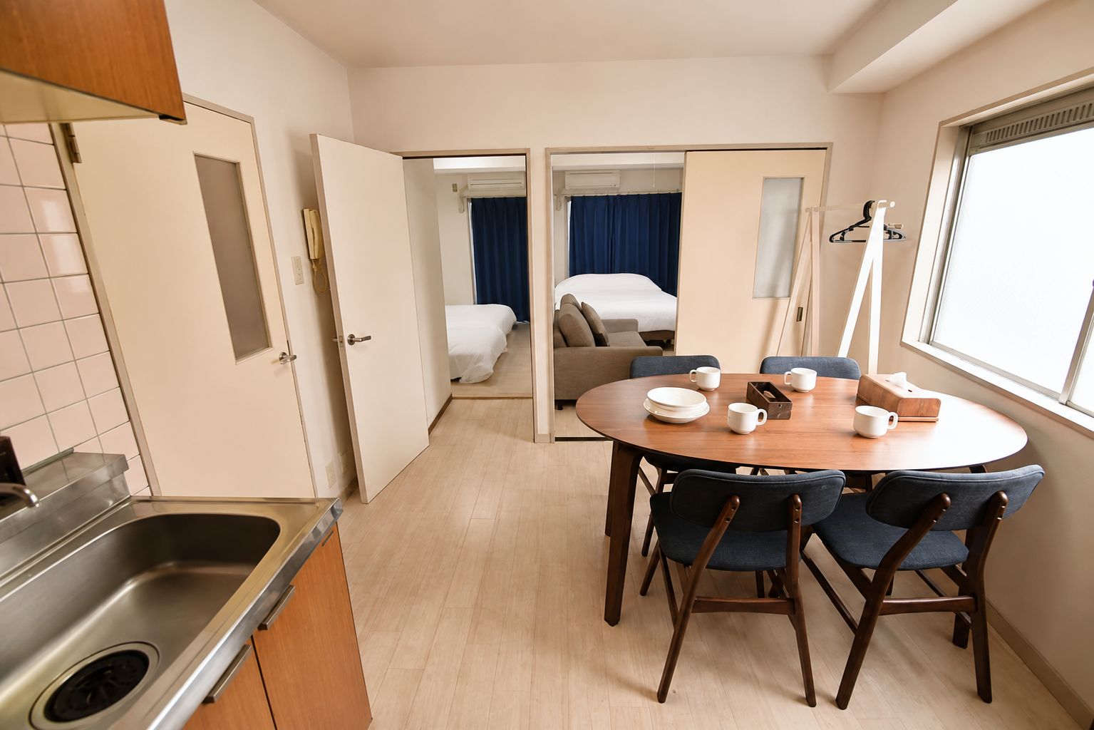 A compact hotel room with a kitchenette, a dining area with a round table and chairs, a bed with a blue curtain, and a large window.