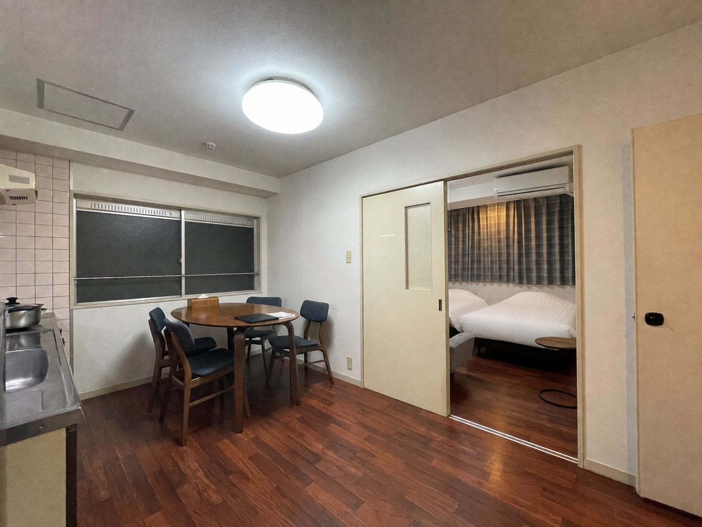 A compact hotel room with a dining area, a kitchenette, and a sliding door leading to a bed.
