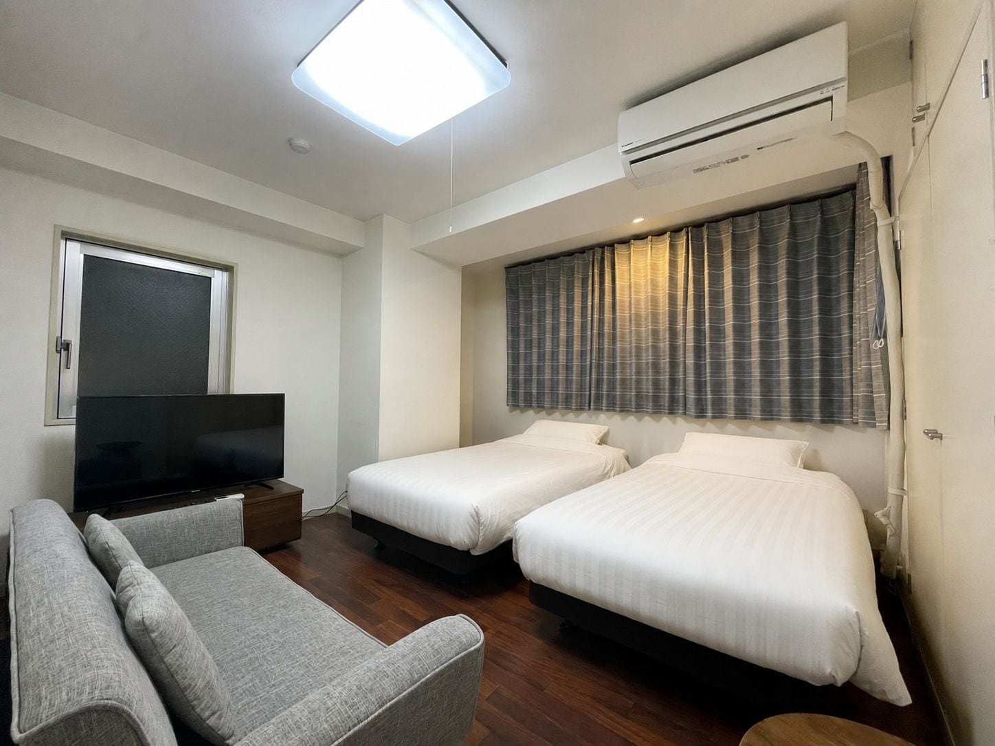 A hotel room with two beds, a sofa, a TV, and an air conditioning unit.