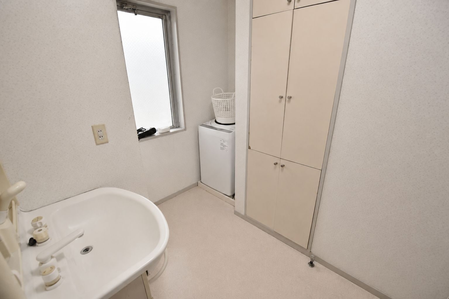 A bathroom with a white sink, a window, a small white laundry machine, and a beige wardrobe.