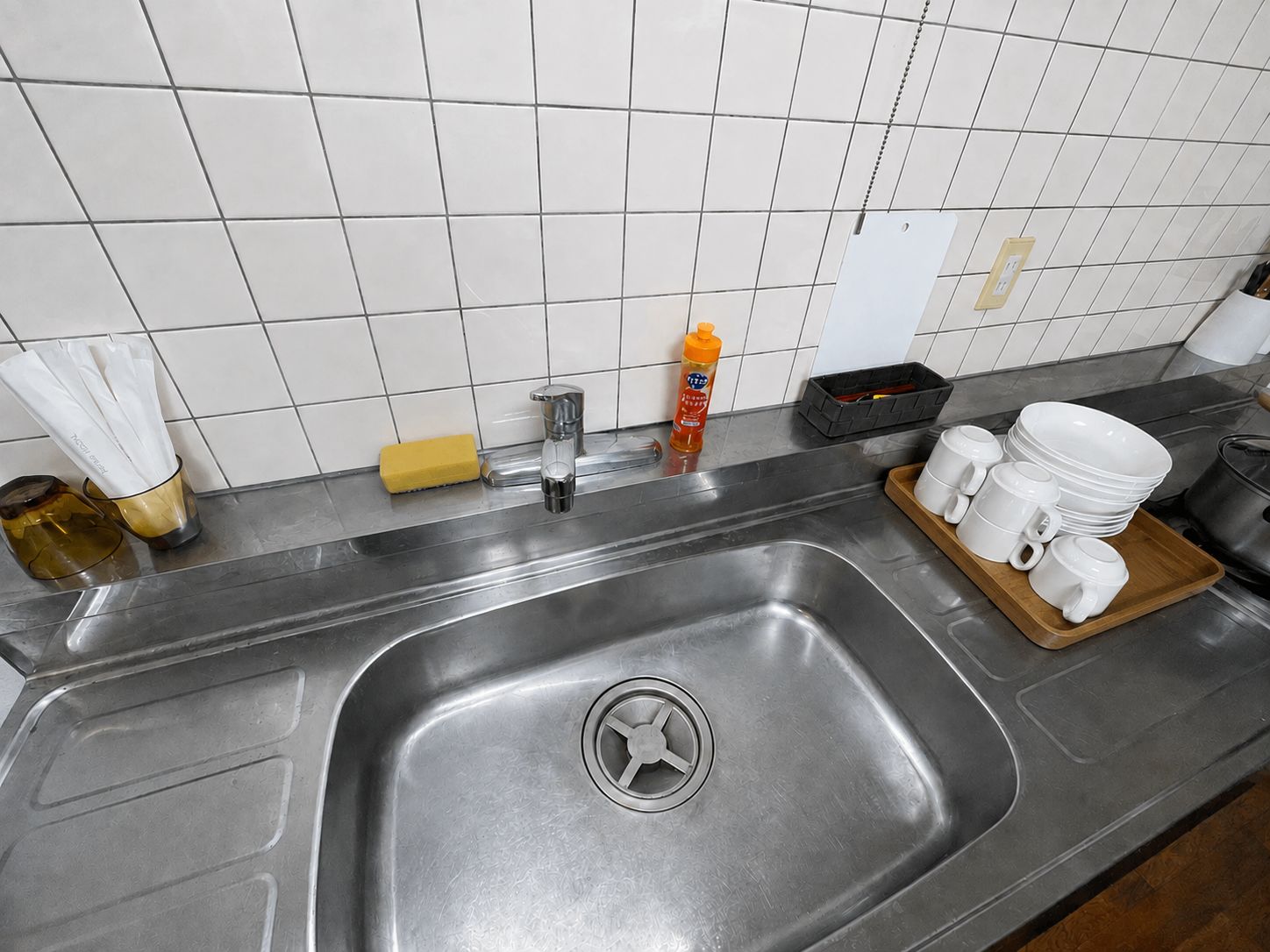 Un lavamanos de acero inoxidable con un solo grifo, un dispensador de jabón y un escurreplatos con tazas y cuencos blancos.