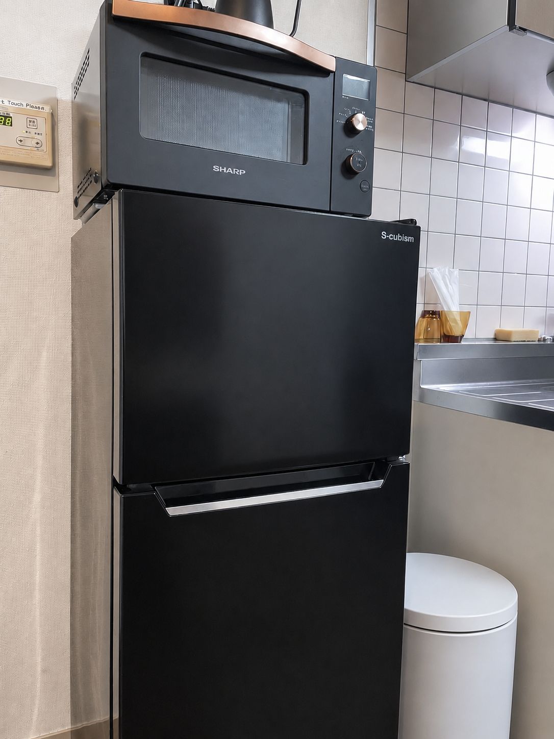 A black Sharp S-cubism refrigerator with a touch panel and a freezer drawer, placed in a kitchen with white tiles and a white trash bin.