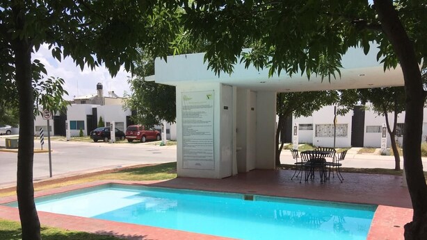 A rectangular pool with a paved edge, surrounded by grass and trees. There is a covered seating area with chairs and a table. A sign with text is visible near the pool. - Hermosa casa completa en coto privado (Aguascalientes)