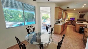 Uma área de refeições com mesa e cadeiras de vidro, uma cozinha com armários de madeira e vista para uma piscina pelas janelas.