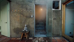 A modern interior with a concrete wall, a small Buddha statue on a wooden table, and a glass door leading to a hallway.