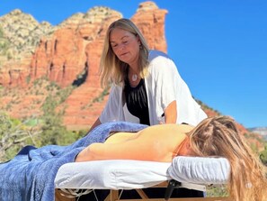 A person lying on a bed with a blanket, another person standing beside them, and a red rock formation in the background. - Eagle Dancer Guest Suite | Wellness Spa & Trails (Sedona)