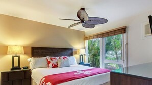 Ein Schlafzimmer mit einem Bett, Nachttischen, Lampen, einem Deckenventilator und einem Fenster mit Blick auf Palmen.