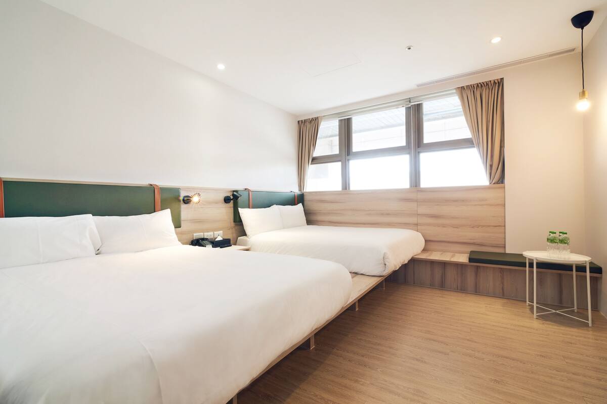 A hotel room with two beds, a small table with bottles, a bench, and a window with curtains.