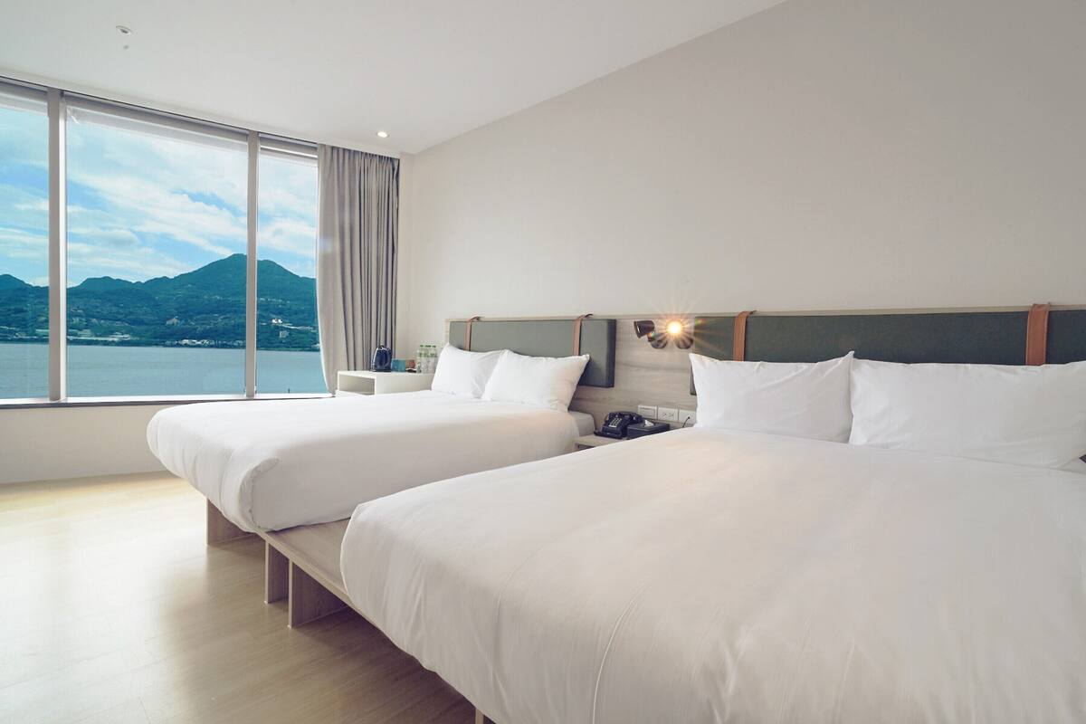 A hotel room with two beds, a large window with a mountain view, and a bedside table with a lamp.