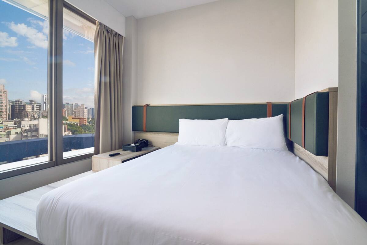 A hotel room with a large bed, a nightstand, a remote control, and a view of the city through a large window.